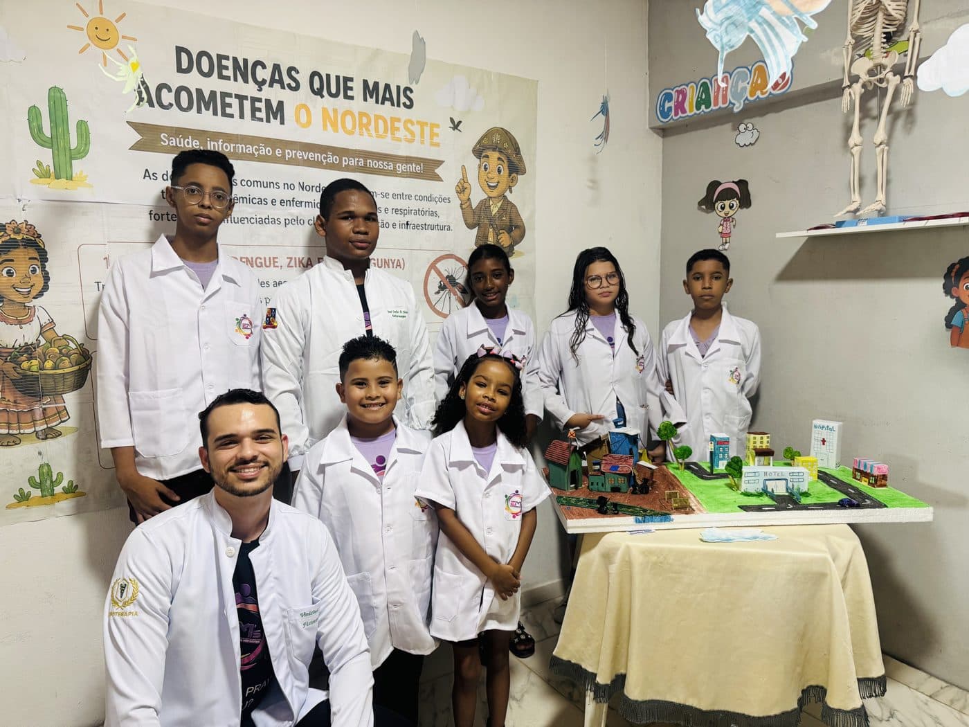 Students in white lab coats with their teacher beside a model city they built