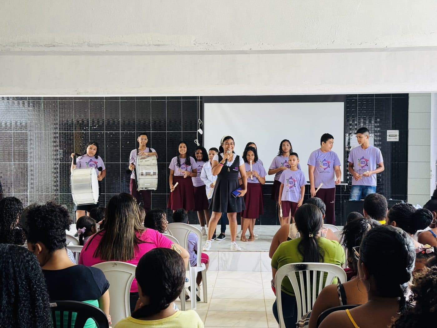 Em with a microphone in front of students playing drums and recorders for their families